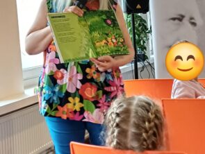 Kobieta w okularach i kolorowej bluzce w kwiaty stoi i czyta dzieciom książkę. W jednej dłoni trzyma mikrofon, a w drugiej pokazuje otwartą, zieloną książkę z ilustracjami przedstawiającymi las i zwierzęta. Przed nią na pomarańczowych krzesłach siedzą słuchające jej dzieci.