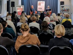 Zdjęcie przedstawia spotkanie autorskie w bibliotece, podczas którego dwie kobiety siedzą na pomarańczowych fotelach na niewielkiej scenie i prowadzą rozmowę. Jedna z nich trzyma mikrofon i zwraca się do drugiej. Za rozmówczyniami znajduje się granatowa kurtyna oraz plakat promujący wydarzenie. Przed sceną. na rzędach czarnych krzeseł, siedzi liczna publiczność, złożona głównie z dorosłych osób, które słuchają rozmowy. Po bokach sceny ustawiono głośniki, a po prawej stronie widoczny jest stojak z książkami. Zdjęcie wykonano z perspektywy widowni, ukazując atmosferę kameralnego spotkania literackiego.