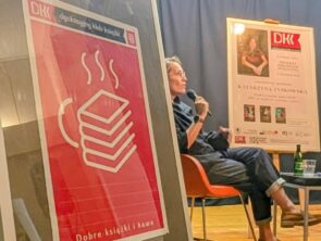 Spotkanie autorskie w ramach Dyskusyjnego Klubu Książki. Na pierwszym planie, po lewej stronie, widoczna jest stojąca sztaluga, a na niej oprawiony w antyramę plakat DKK. Znajduje się na nim grafika przedstawiająca filiżankę w kształcie stosu książek, nad którą unosi się para. Poniżej znajduje się napis „Dobre książki i kawa". W głębi, na drewnianej scenie, na pomarańczowym nowoczesnym krześle-fotelu, siedzi kobieta w ciemnym ubraniu i okularach. trzyma w dłoni mikrofon i przemawia do publiczności.